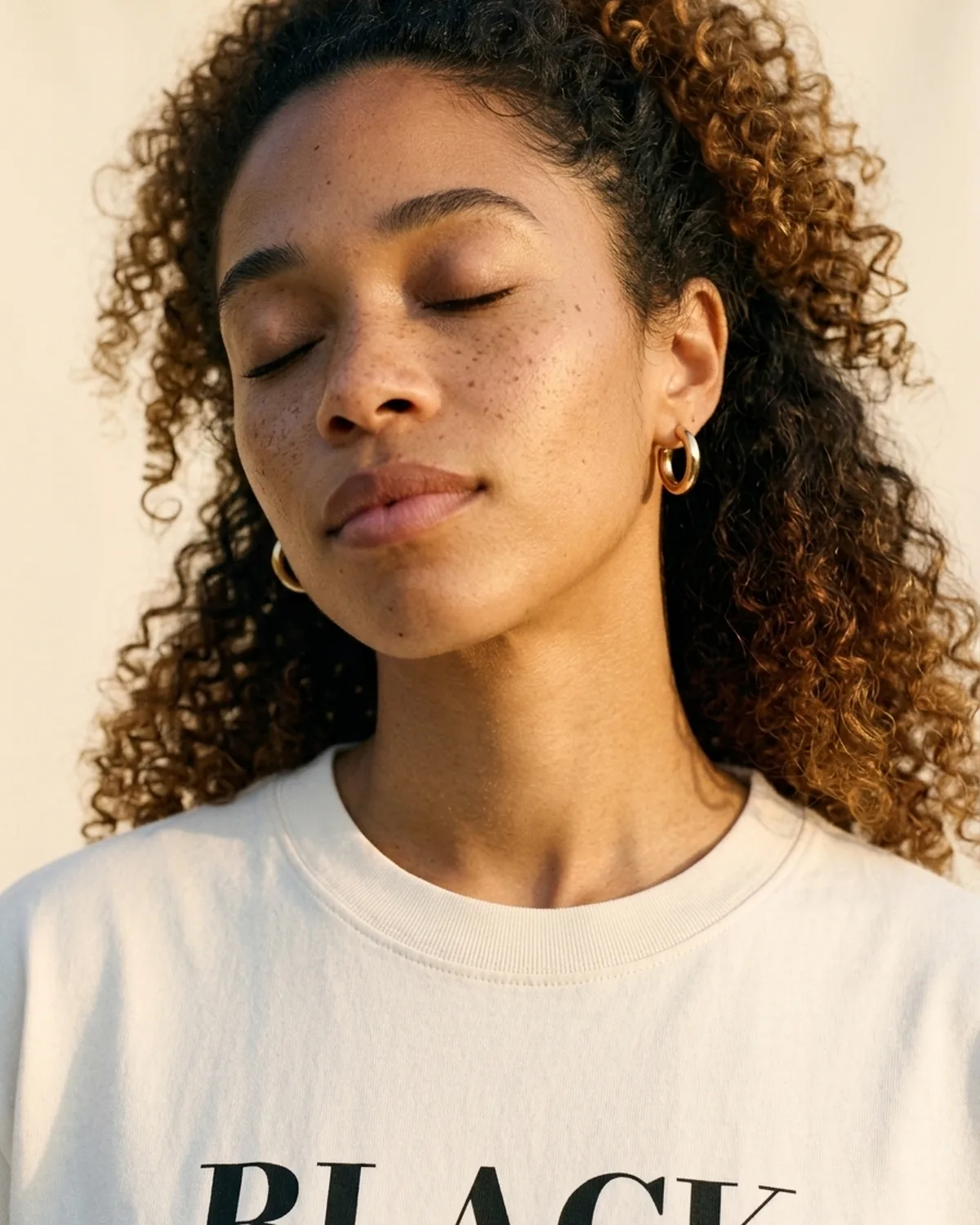 Nia with eyes closed and head tilted, warm light on her neck and collarbone, wearing the Ivory Whole tee
